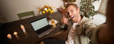 Yakışıklı genç adam çalışırken ya da laptopla uzaktan çalışırken selfie çekiyor, telefon işaretini gösteriyor ve cep telefonu kamerasına gülümsüyor..