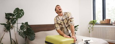 Giysilerle dolu bavulu kapatmaya çalışan bir adamın portresi, bayram için bavul hazırlama çabası, uzun bir tatile çıkma. Turizm ve seyahat acentası kavramı