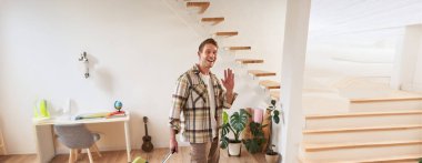 Bavullu mutlu bir gencin dikey görüntüsü, kiralık daireye giriş yapıyor, kameraya el sallıyor, içeride bavulla yürüyor..