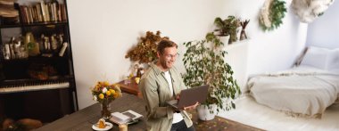 Yakışıklı, mutlu bir adamın dikey portresi, şirketin yöneticisi veya CEO 'su, masada oturuyor ve dizüstü bilgisayarını tutuyor, gülüyor, görüntülü sohbet ediyor, bir ekiple çevrimiçi bir toplantı yapıyor, ofiste duruyor..