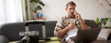 Kafası karışmış bir adam, cep telefonuyla konuşuyor, dizüstü bilgisayarla oturuyor, yanına bavulu yerleştiriyor, seyahat acentasıyla tatille ilgili sorular soruyor..