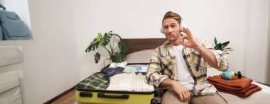 Yakışıklı sarışın adamın portresi, turist tatile çıkıyor, kablosuz kulaklıkla müzik dinliyor, kıyafetlerini bavula koyuyor, tatil yerini seçiyor, iyi el hareketi gösteriyor..