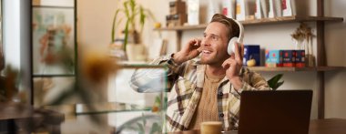 Gülümseyen dijital göçebenin yaşam tarzı portresi, kafede çalışan adam, dizüstü bilgisayarda çalışan, kablosuz kulaklıkla müzik dinleyen, gülümseyen, sandalyede dans eden ve favori şarkının keyfini çıkaran..