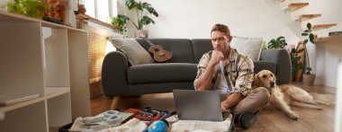 Genç bir adamın resmi, tatile çıkan bir turist, oturma odasında bir köpekle oturuyor, dizüstü bilgisayara bakıyor, bilgisayardaki bir uçağa giriş yapıyor. Turizm ve seyahat kavramı