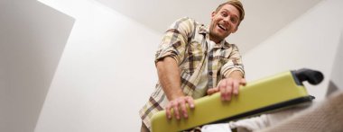 Komik mutlu adamın düşük açılı çekimi, bavulunu toplayan turist, giysi dolu çantasını kapatmaya çalışıyor. Turizm ve seyahat kavramı