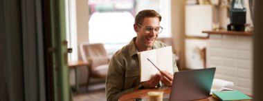 Gözlüklü genç yakışıklı bir adamın portresi, özel ders veren bir öğretmen, defterine işaret eden, bir şeyler gösteren, laptop uygulaması üzerinden video sohbetleri, kafede uzaktan çalışan.