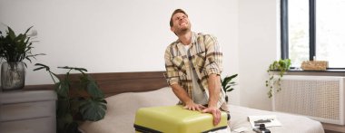 Terleyen genç adamın portresi, bavulunu kapatması, bir sürü kıyafetle valizi kapatmaya çalışması, yatağa oturması, yaz tatili için eşyalarını toplaması, ya da iş seyahatine çıkması..