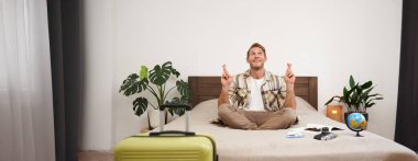 Yaz tatili için bavulunu topladıktan sonra yatakta meditasyon yapan genç bir adamın portresi, bacak bacak bacak üstüne atmış, nirvana pozu veren, rahatlatıcı, bir şeyler umut eden. Turizm ve farkındalık kavramı