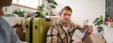 Elinde bavuluyla gülümseyen yakışıklı bir adamın portresi, akıllı telefona bakıyor, seyahat bileti ve pasaport tutuyor. Turizm ve seyahat kavramı