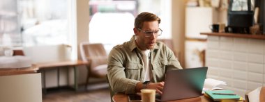 Ciddi yüzlü bir adamın portresi, kafede oturuyor, gözlük takıyor, dizüstü bilgisayarda çalışıyor, konsantre görünüyor, serbest çalışıyor..