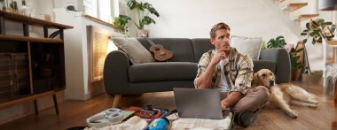 Genç bir adamın portresi, gezgin bir köpekle tatile çıkıyor, bavulunu topluyor, laptopuyla oturuyor, düşünüyor, online rezervasyon yapıyor..