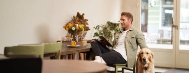 Yakışıklı genç adamın portresi köpeğiyle kafede oturuyor Golden Retriever 'ı okşuyor, tabletten haberler okuyor, bir fincan kahveyle rahatlıyor..