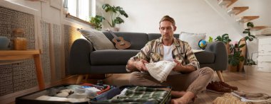 Tatil için kıyafet seçen genç bir adamın portresi, bavulla yerde oturmak, tatile çıkmak, iş seyahatine çıkmak..