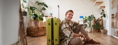 Gülümseyen yakışıklı adam pasaportuna ve uçuş biletine bakar, bavuluyla yere oturur..