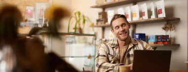 Yakışıklı genç dijital göçebe portresi, kafede dizüstü bilgisayarla çalışan, online projesinden memnun ve mutlu görünen, birlikte çalıştığı kahve dükkanında oturan bir adam..