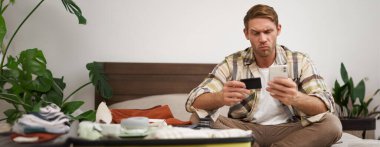 Düşünceli yüzlü bir adamın portresi, bavulla yatakta oturuyor, internetten kredi kartı ve akıllı telefon uygulaması ile ödeme yapıyor. Boşluğu kopyala