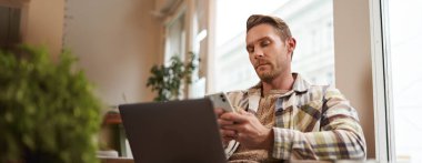 Ciddi yüzlü bir adamın portresi, cep telefonuyla haber yayını, akıllı telefon ekranına odaklanmış, kahve ve dizüstü bilgisayarla kafede oturuyor..