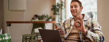 Adamın olumlu görüşlerini yansıtan portresi, dizüstü bilgisayarla otururken, tamam, tamam jestini gösteriyor. Cafe ziyaretçisi cep telefonu bulundurarak iyi bir çalışma alanını onayladı.