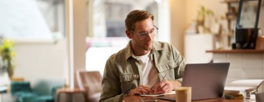 Gözlüklü, kafede oturan, notlar alan, çevrimiçi kurslara katılan, uzaktan kumandalı bir kafeden öğrenen odaklanmış bir genç resmi..