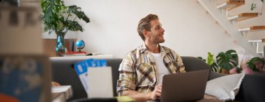 Yakışıklı bir gencin portresi, iki bileti ve pasaportu var, dizüstü bilgisayara bağlanıyor, otel rezervasyonu yapıyor ve tatilini planlıyor, oturma odasında oturuyor..