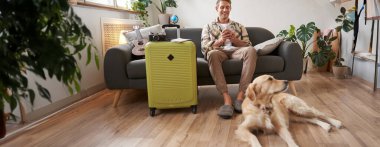 Bavullu, mutlu, yakışıklı genç bir adamın portresi, köpeğiyle oturma odasında oturuyor, tatile çıkmaya hazırlanıyor, tatile çıkıyor. Turizm konsepti