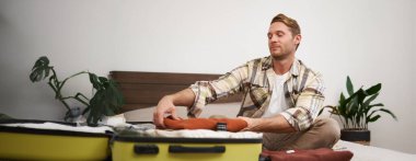 Yakışıklı genç bir adamın portresi, otel odasında bavulları boşaltan turist, tatilde pansiyonda kalan, seyahat eden, yatakta bavullarla oturan..