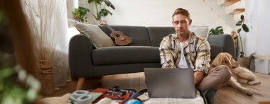 Bavulunda dizüstü bilgisayarı olan bir adam, yerde köpekle oturuyor, internete bakıyor, web sitelerine bakıyor, tatil için kıyafet paketliyor, tatile çıkıyor, planlar yapıyor..