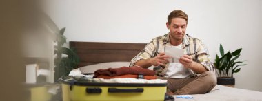 Yakışıklı bir adamın portresi, turist, tatil biletlerine bakıyor seyahat acentesinde yer ayırtıyor, yatakta oturuyor ve giysilerini bavuluna koyuyor..