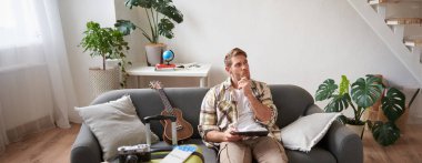 Tabletli genç bir adamın resmi, oturma odasında bavuluyla oturuyor, okuyor, rotayı okuyor, tatile hazırlanıyor. Turizm konsepti