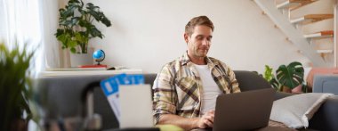 Yakışıklı bir gencin portresi, iki bileti ve pasaportu var, dizüstü bilgisayara bağlanıyor, otel rezervasyonu yapıyor ve tatilini planlıyor, oturma odasında oturuyor..