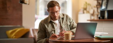 Gözlüklü, kafede oturan, notlar alan, çevrimiçi kurslara katılan, uzaktan kumandalı bir kafeden öğrenen odaklanmış bir genç resmi..