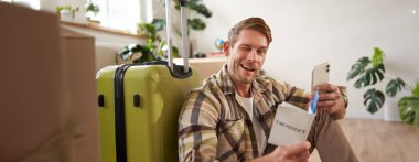 Karizmatik genç adam, tatile çıkan yolcu, yurtdışına seyahat, bavulla oturan, akıllı telefon, bilet ve pasaport tutan, kameraya göz kırpan ve gülümseyen.