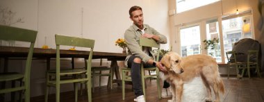 Kahve dükkanında siparişini bekleyen, bir köpeği okşayan, hayvan dostu bir kafede dinlenen yakışıklı genç adamın yakın portresi..