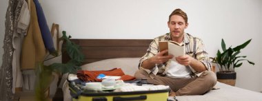 Bavullu bir adamın portresi, tatil için otururken ve eşyalarını toplarken, tatil için eşya listesini kontrol ederken, not alırken, not defterine not alırken. Yaşam tarzı ve turizm konsepti