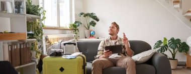 Tabletli heyecanlı bir genç, valiziyle oturma odasında oturuyor, şans dilemek için dua ediyor, dilek tutuyor, bir şeyler umut ediyor. Seyahat konsepti