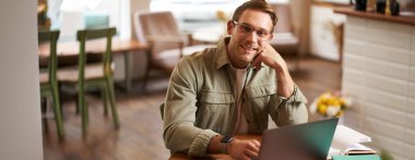 Uzaktan çalışma ve serbest çalışma kavramı. Gözlüklü yakışıklı adam dizüstü bilgisayar ve dizüstü bilgisayarla oturur, online çalışır, kahve dükkanından uzaktan çalışır, bilgisayarla kafede oturur..