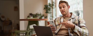 Serbest çalışan genç bir adamın portresi, kafede çalışıyor, dizüstü bilgisayarla sandalyede oturuyor, telefon ekranını işaret ediyor, telefonundaki bir şeyle ilgileniyor..