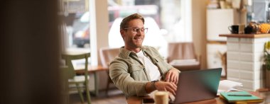 Kafede dizüstü bilgisayarla çalışan, kendinden memnun ve memnun görünen, bilgisayar ekranı için gözlük takan bir adamın portresi, kahve dükkanında sandalyeye oturup.