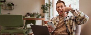 Yakışıklı erkek kafe ziyaretçisinin portresi, dizüstü bilgisayarda çalışan iş adamı, akıllı telefon tutan, cep telefonu işareti gösteren, beni ara..