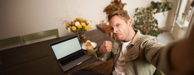 Yakışıklı bir adamın portresi, Selfie fotoğrafı için kızgın tehditkar yüz ifadesi, kafede akıllı telefon uygulamasında fotoğraf çekmek, dizüstü bilgisayarın yanında oturmak..