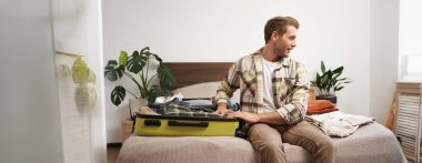 Turizm ve seyahat konsepti. Yakışıklı genç adamın portresi tatile çıkıyor, bavulunu topluyor, bavullarını kontrol ediyor, yatağa oturuyor ve paketlenmiş eşyalara bakıyor..