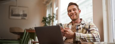 Gülümseyen, kahve dükkanında dinlenen yakışıklı genç bir adamın portresi. Kafede ortak çalışma alanından bilgisayarı ve akıllı telefonu kullanarak çalışıyor ve kameraya bakıyor..