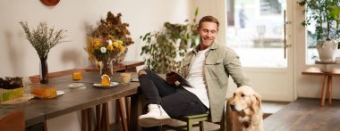 Mutlu gülümseyen genç bir adamın portresi, kafe ziyaretçisi, köpeğiyle kahve dükkanında oturuyor, Golden Retriever 'ı okşuyor, dijital tablet tutuyor, haberleri okuyor ya da internette geziniyor..