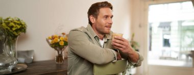 Yakışıklı bir adamın yakın portresi, sandalyede oturuyor, elinde bir bardak kahve, cappuccino içiyor ve gülümsüyor. Yaşam tarzı ve eğlence anlayışı.