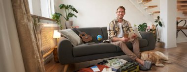 Elinde kontrol listesi olan genç bir adamın portresi. Köpeğiyle tatile çıkıyor. Tatil için bavul hazırlıyor..