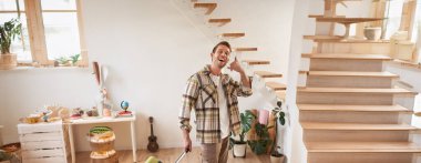 Tatile çıkan mutlu genç adamın dikey portresi, cep telefonu el hareketi, bavulla yürümek, kiralık daireden çıkmak. Seyahat ve turizm konsepti