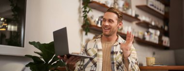 Kafedeki genç bir adamın görüntüsü, video görüşmesine bağlanıyor, çevrimiçi konuşuyor, bilgisayarla arkadaşlarıyla sohbet ediyor ve kameraya selam vermek için el sallıyor..