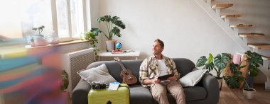 Yakışıklı genç adamın portresi, bavul ve iki bilet yanında dijital tabletle oturan turist, tatili için internetten bir şeyler ayırtıyor, tatil hazırlıyor..