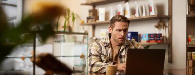 Genç bir işletme sahibi, kafeteryada dizüstü bilgisayarla oturan kafası karışık bir şekilde ekrana bakan dijital göçebe işi çözmeye çalışan bir programcı..