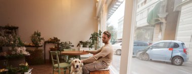 Golden Retriever 'ın sahibi genç bir adamın dikey pozu. Kafede köpeğiyle birlikte masanın altında oturup kahve içerken..